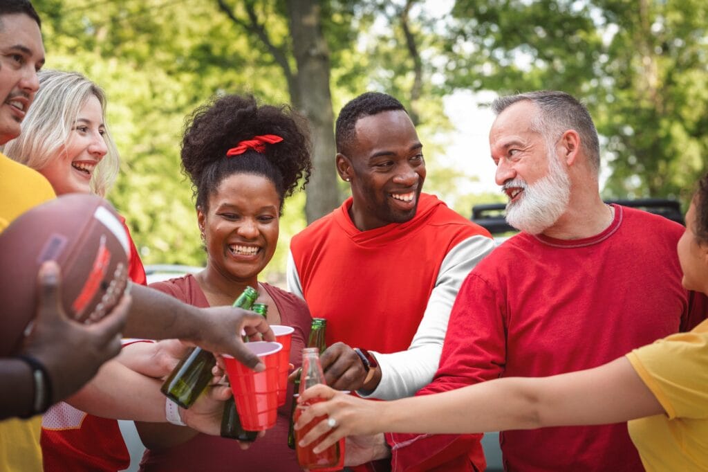 A diverse group of friends laugh and toast with drinks at an outdoor gathering, sharing a lighthearted moment that reflects the spirit of "read the room" social awareness.