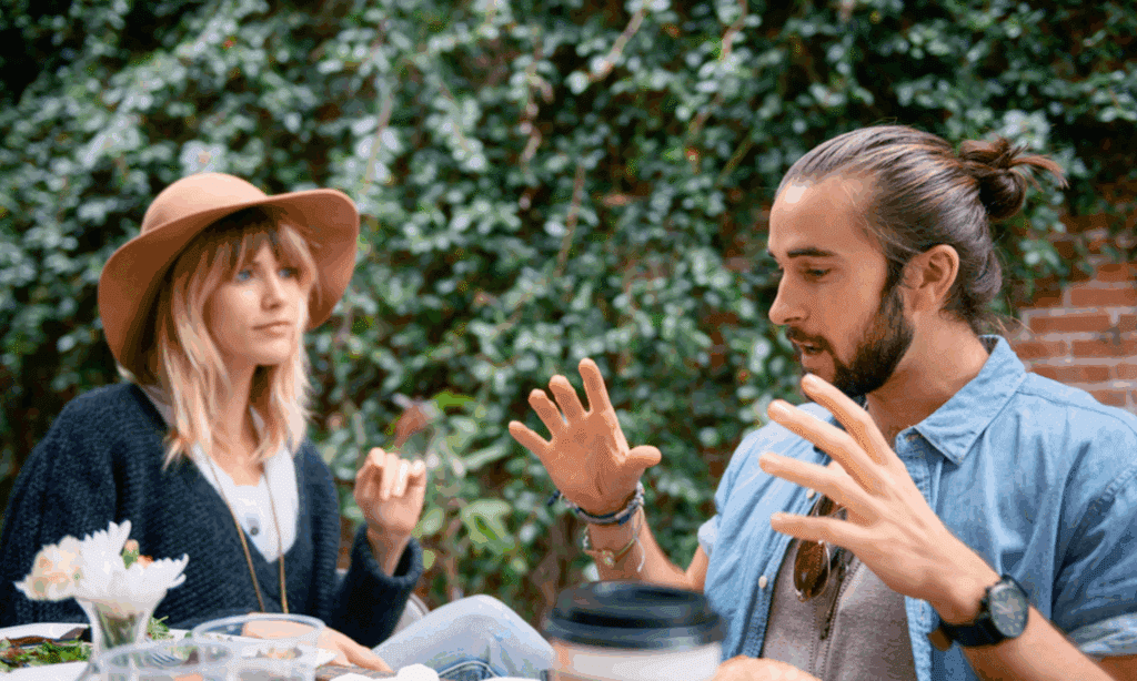 Figure 1. Uneven engagement at an outdoor café. A man and woman sit across a small table framed by ivy-covered brickwork. The man leans forward, both hands lifted above the tabletop with fingers spread in a wide, animated gesture. His open palms, raised shoulders, and slightly parted lips signal high arousal and a strong drive to dominate the conversational floor. By contrast, the woman’s torso angles away from him. She reclines against the bench, eyes drifting past his face rather than locking on it, her mouth closed in a neutral line. One hand hovers at chest height in a loose pinch. It looks as if it is poised to respond but never quite joining in. While the other rests on her lap. The asymmetry between his expansive gestures and her minimal, withdrawn posture, coupled with her indirect gaze (notice how she isn't looking at him but at what's behind him), suggests she is only half-engaged and that he has not yet “read the room” to notice her fading interest.