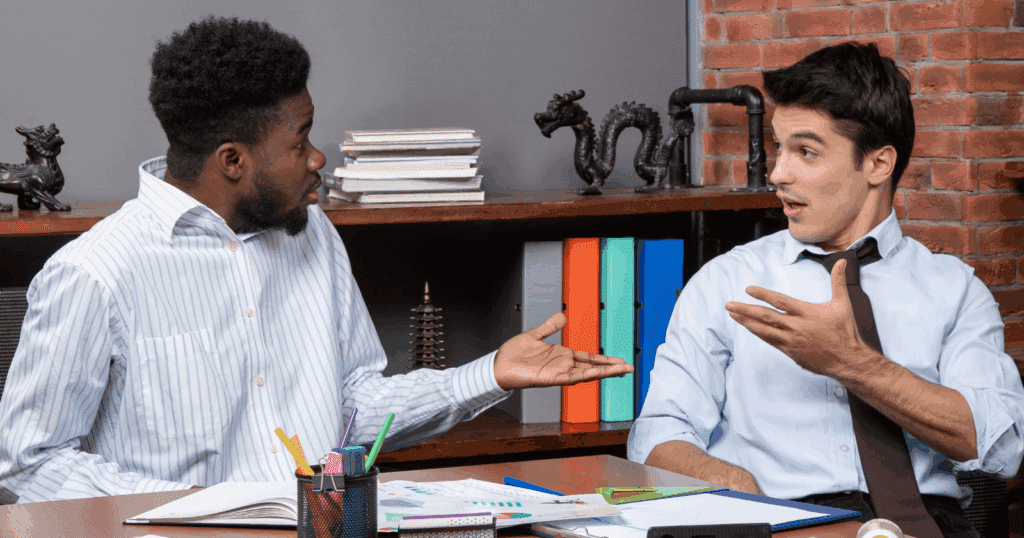 Figure 2. Mutual surprise and implicit challenge in an office exchange. Two colleagues face one another across a cluttered desk, each frozen mid-gesture. The man on the left angles his torso toward the other, eyebrows lifted and eyes wide; his upturned palm and slightly parted lips wordlessly ask “seriously? ” or “explain yourself. ” the man on the right mirrors the widened eyes and open mouth, but his body tilts back, tie loosened, and his pointing hand redirects the spotlight: “wait—me? ” or “hold on, you did that. ” their parallel expressions (raised brows, slack jaws), open palms, and synchronous hand-motions create a rapid feedback loop of shared astonishment and low-grade confrontation. No one is smiling, and both are leaning away from the desk rather than toward collaboration. These are non-verbal cues that signal surprise, mild blame, and a momentary breakdown in cooperative rapport.