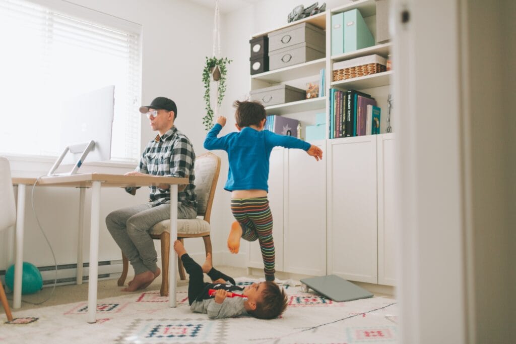 A trad husband works from home at a desk while his young children play energetically around him, showing the multitasking and family dynamics of modern traditional fatherhood.
