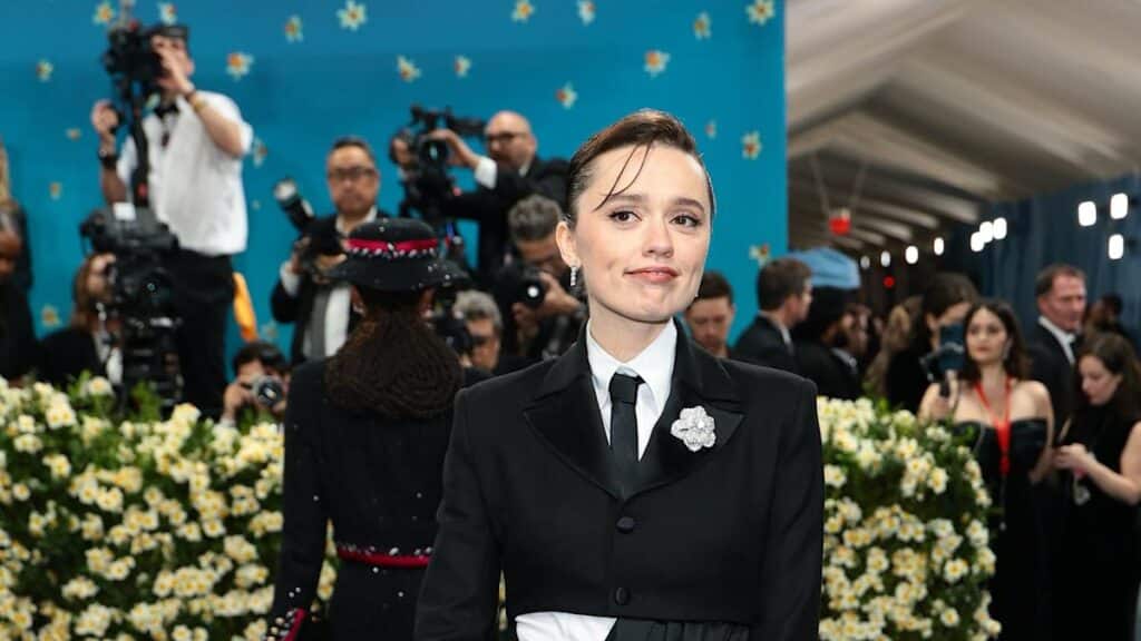 Aimee Lou Wood at the 2025 Met Gala wearing a black tuxedo-style gown with a white flower brooch, standing in front of a floral backdrop and a crowd of photographers.