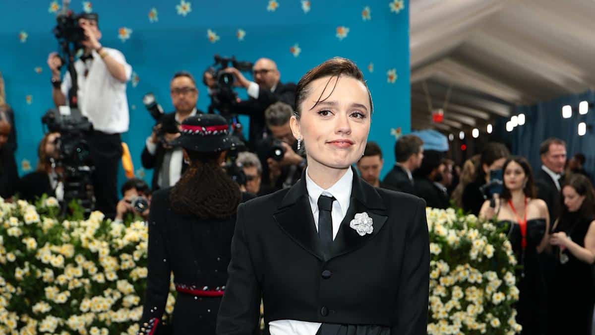 Aimee Lou Wood at the 2025 Met Gala wearing a black tuxedo-style gown with a white flower brooch, standing in front of a floral backdrop and a crowd of photographers.