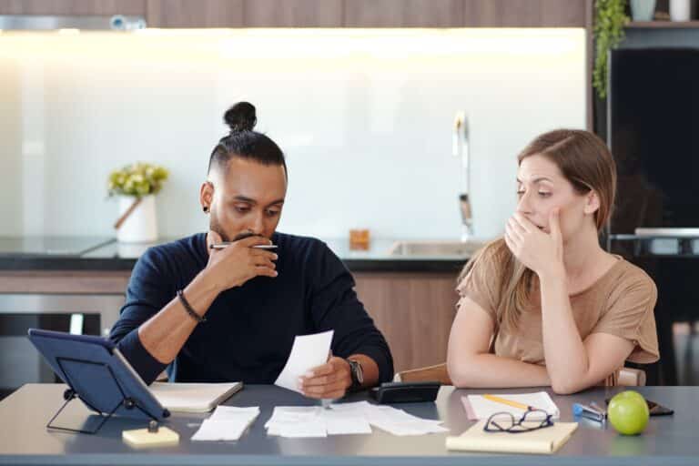 A man and woman sit at a kitchen table, visibly distressed while reviewing financial documents. The man looks down at a receipt, pen in hand, and the woman covers her mouth in concern. Papers, a tablet, and notebooks are spread across the table, indicating a serious discussion about debt.