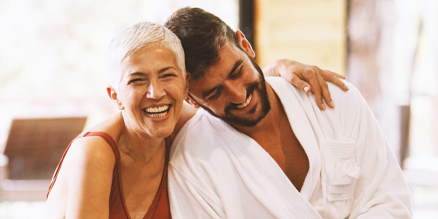 Smiling older woman with short gray hair and a younger bearded man sitting closely together, showing affection and happiness.