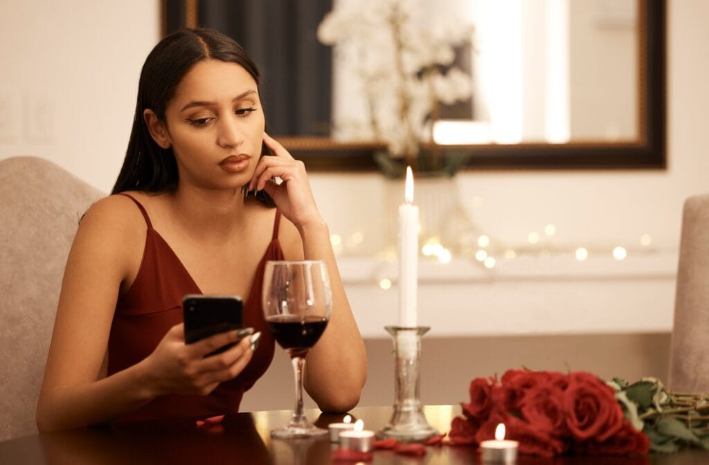 A woman in a red dress sits alone at a romantic dinner table, looking at her phone with a disappointed expression, suggesting she is still single.