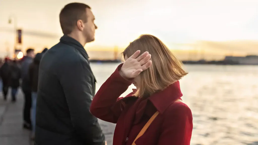 A woman in a red coat raises her hand to block her face while standing beside a man near the water, symbolizing common triggers for fearful avoidant partners.
